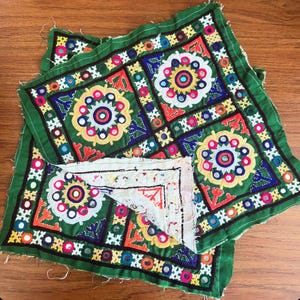 May include: A square green fabric piece featuring intricate embroidery in yellow, red, blue, and white, accented with small circular mirrors. The design includes geometric patterns and floral motifs, with a partially folded corner revealing a white lining.