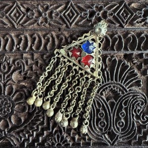 May include: A vintage brass pendant with a triangular design, featuring red and blue glass inserts. The pendant is adorned with dangling chains and small bell charms, set against a dark, carved wooden surface. A unique piece of jewelry.