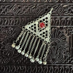 May include: Silver-toned triangular pendant with a red gemstone center and dangling chains. The pendant has a filigree design and small bell-shaped charms at the chain ends. A vintage-style piece.