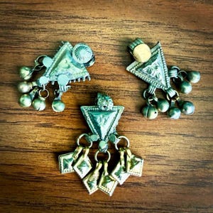 May include: Three triangular metal pendants with a weathered patina. Each piece features a central triangle with dangling charms, including small spheres and diamond-shaped plates attached by metal rings. The metal has a muted, aged appearance.