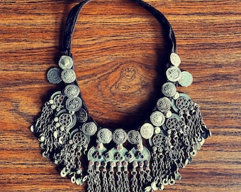 May include: A silver necklace with a large pendant made of multiple round silver coins with intricate designs. The pendant is surrounded by smaller silver coins and chains. The necklace is on a brown wooden surface.