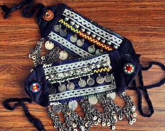 May include: Two blue fabric bracelets with silver coin accents, white and green beads, and colorful embroidery. The bracelets are tied together with a black cord and have silver chain tassels.