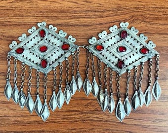 May include: Silver-toned metal jewelry with red gemstone accents. The design features a geometric shape with dangling leaf-shaped pendants. The jewelry is displayed on a wooden surface, showcasing its intricate details and craftsmanship.