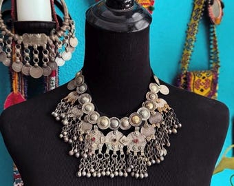 May include: Silver-toned statement necklace with coin and chain details. The necklace features multiple circular and geometric elements, with small red accents. It is displayed on a black mannequin, showcasing its intricate design.