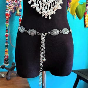 May include: Silver-toned metal belt with oval-shaped decorative elements and a chain tassel. The belt is displayed on a black mannequin, showcasing its design and style. The belt is a fashion accessory.