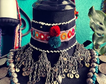 May include: A black mannequin wearing a silver chain necklace with multiple silver coin charms. The necklace has a black and white beaded band with colorful embroidery and a red and green pom-pom.