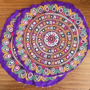 May include: Two circular purple fabric mats featuring intricate embroidery in yellow, green, and white. The design includes concentric rings of geometric patterns, floral motifs, and small reflective circular mirrors embedded throughout the surface.