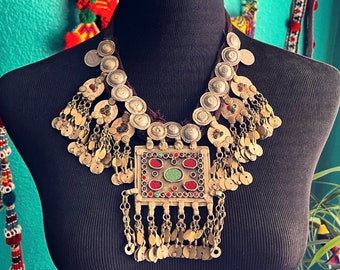 May include: A silver and gold necklace with a large, ornate pendant featuring red, green, and blue stones. The necklace is made of multiple layers of beads and chains, and has a tassel-like fringe.