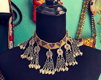 May include: A beaded choker necklace with a yellow and purple pattern and silver pendant charms. The necklace is displayed on a black mannequin.