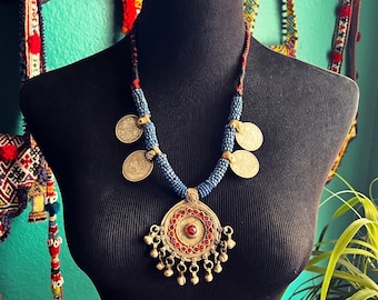 May include: A beaded necklace with a large silver pendant and coin accents. The pendant has a red center and dangling bells. The necklace is displayed on a black mannequin.