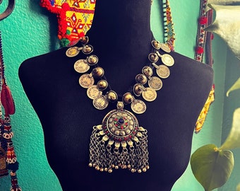 May include: A silver necklace with a large, round pendant featuring a multi-colored stone surrounded by smaller stones. The pendant is attached to a chain of silver coins with a black cord.