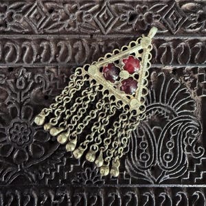 May include: A silver-toned triangular pendant with three red gemstones and dangling chains. The pendant has a filigree design and small gold-colored bell-shaped charms at the chain ends. The background is a dark, carved wooden surface.