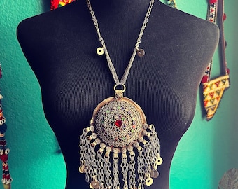 May include: A silver statement necklace with a large, ornate pendant. The pendant features a central red stone surrounded by smaller colourful gems and intricate metalwork. The necklace has a chain with small coin-like charms and a fringe of dangling chains.