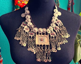 May include: A large, ornate silver necklace with a rectangular pendant featuring red and green stones. The necklace is made of multiple layers of round silver discs and chains with small silver discs hanging from them.
