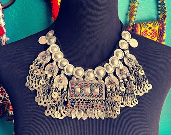 May include: A silver necklace with a large, ornate pendant featuring colorful gemstones. The pendant is surrounded by smaller silver beads and chains.
