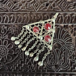 May include: Silver-toned triangular pendant with three red gemstones and dangling chains. The pendant has a filigree design and small silver beads at the end of each chain. The background is a dark, carved wooden surface.