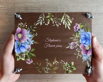 May include: A wooden flower press with a floral design in shades of blue, purple, and green. The press has metal hardware and the text "Stephanie's Flower press" is visible. The press is held by two hands.