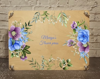 May include: A wooden flower press with a floral design featuring blue and purple flowers. The text "Mariya's Flower Press" is printed on the front.