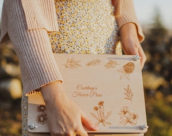 May include: A wooden flower press with the text "Courtney's Flower Press" engraved on the front. The press features a variety of floral designs, including leaves, flowers, and a clover.