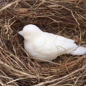 May include: A white bird figurine rests in a nest made of twigs and dried grass. The bird is smooth and solid, with a simple, elegant design. The nest is a natural brown color, providing a rustic contrast to the bird's pristine white.