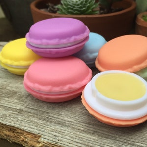 May include: Six colorful, round, macaron-shaped lip balm containers. The containers are in shades of yellow, pink, purple, blue, and orange. The lip balm is visible in one container, which is a light yellow color.