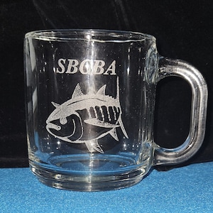 May include: A clear glass mug with a handle, featuring an etched design of a tuna fish and the text "SBQBA".