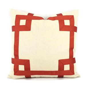 May include: A cream-colored pillow with a red ribbon border. The ribbon is arranged in a geometric pattern, creating a square frame around the pillow.