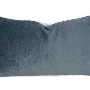 Puede incluir: Un coj&iacute;n rectangular con una textura suave similar al terciopelo en un color azul gris&aacute;ceo profundo y apagado. La tela muestra un brillo sutil en toda su superficie, con una ligera hendidura en el centro superior, lo que le da un aspecto mullido.