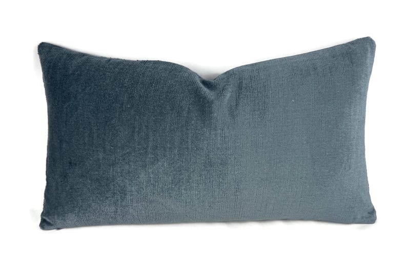 Puede incluir: Un coj&iacute;n rectangular con una textura suave similar al terciopelo en un color azul gris&aacute;ceo profundo y apagado. La tela muestra un brillo sutil en toda su superficie, con una ligera hendidura en el centro superior, lo que le da un aspecto mullido.
