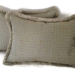 May include: Two decorative throw pillows with a geometric pattern and fringe trim. The pillows are a light green color with a subtle gold thread design.