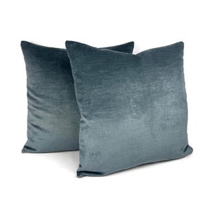 May include: Two square throw pillows featuring a solid, deep blue-grey velvet fabric. The material has a soft, light-reflecting texture with a consistent color throughout. The pillows are positioned slightly overlapping against a plain white background.