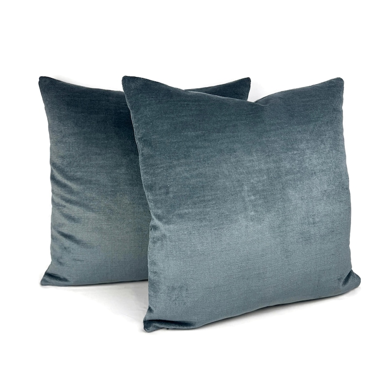 May include: Two square throw pillows featuring a solid, deep blue-grey velvet fabric. The material has a soft, light-reflecting texture with a consistent color throughout. The pillows are positioned slightly overlapping against a plain white background.
