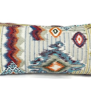 May include: Rectangular decorative pillow with a colorful geometric pattern. The pillow features a mix of blue, red, orange, and yellow zigzags and diamond shapes on a cream background. The pillow is approximately 24 inches long.