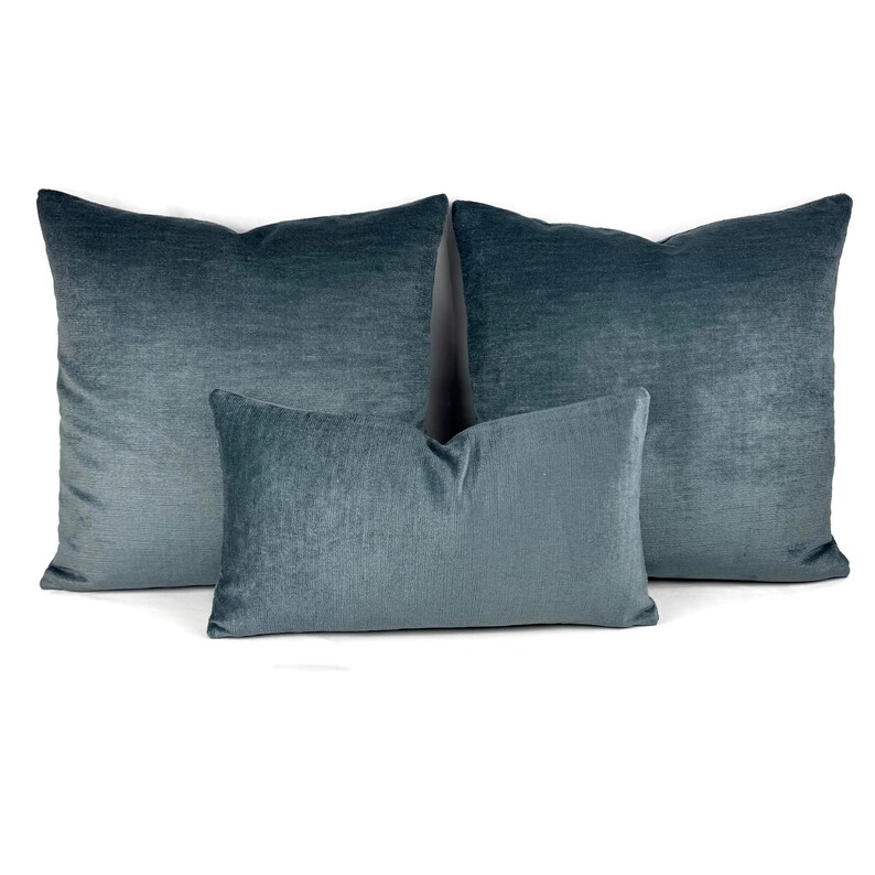 Puede incluir: Tres cojines decorativos cuadrados y rectangulares en un color azul cerceta oscuro s&oacute;lido. Los cojines presentan una textura de tela suave similar al terciopelo con un brillo sutil que refleja la luz en la superficie de cada coj&iacute;n de forma uniforme.