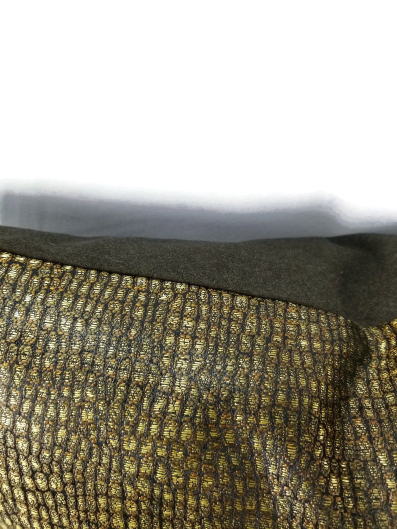 May include: Close-up of a section of fabric with a textured, gold and black pattern. The fabric has a woven appearance with a repeating geometric design. A dark green, solid-colored fabric is visible at the top of the image.