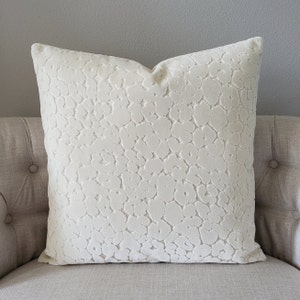 May include: A white velvet pillow with an embossed, abstract pattern. The pillow is on a light beige couch.