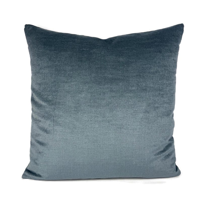 May include: A square throw pillow featuring a soft, velvet-like fabric in a deep blue-grey tone. The material displays a subtle horizontal texture and a slight sheen, creating a smooth, uniform appearance across the entire surface of the cushion.