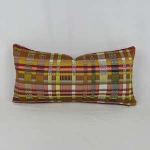 May include: A rectangular throw pillow featuring a woven plaid pattern in shades of mustard yellow, olive green, bright lime, white, and muted red. The fabric displays a complex grid of intersecting horizontal and vertical stripes across the surface.