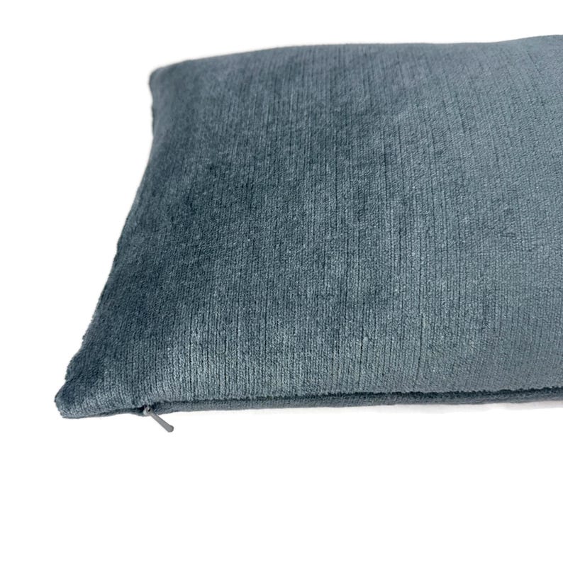Puede incluir: Una funda de coj&iacute;n rectangular de color azul pizarra s&oacute;lido. La tela presenta una superficie suave y texturizada con rayas tipo pana. Una peque&ntilde;a cremallera azul a juego es visible a lo largo del borde inferior para facilitar el cierre.