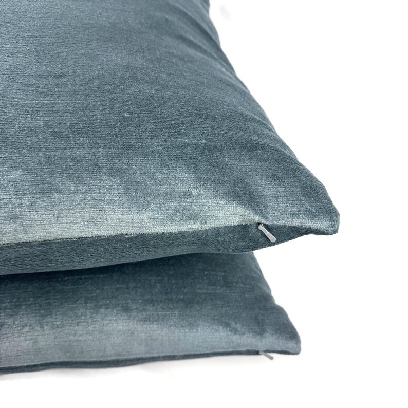 May include: Two stacked square throw pillows in a deep teal blue velvet fabric. Each pillow features a soft, short-pile texture and a discreet, matching zipper closure located along the side seam, providing a clean and uniform appearance.