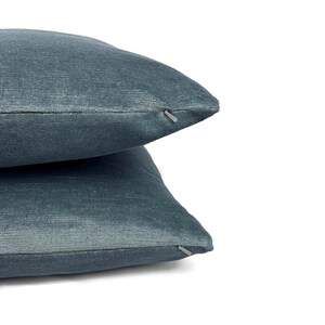 May include: Two stacked square throw pillows in a solid slate blue color. The fabric has a subtle, fine-textured horizontal weave. Each pillow features a small, matching blue zipper closure visible along the side seam, providing a clean finish.