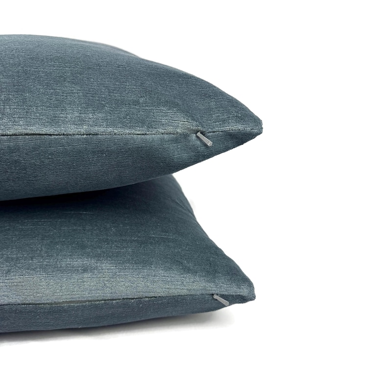 May include: Two stacked square throw pillows in a solid slate blue color. The fabric has a subtle, fine-textured horizontal weave. Each pillow features a small, matching blue zipper closure visible along the side seam, providing a clean finish.