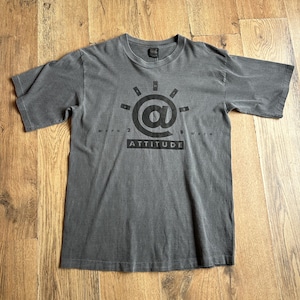 May include: A charcoal grey short-sleeve t-shirt featuring a black graphic on the chest. The design includes an at symbol surrounded by small rectangles, with the text "WORN 2 B WORN" and the word "ATTITUDE" printed in a black rectangular box.