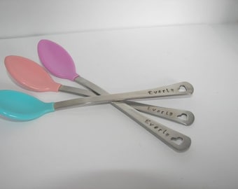 May include: Three stainless steel spoons with heart-shaped cutouts at the end of the handle. Each spoon has a different colored silicone tip: pink, light blue, and peach. The spoons are engraved with the name "Everly".
