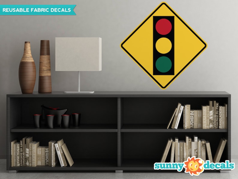 K&ouml;nnte beinhalten: Ein gelbes, rautenf&ouml;rmiges Verkehrszeichen mit einer roten, gelben und gr&uuml;nen Ampel darin. Das Schild tr&auml;gt oben die Aufschrift "REUSABLE FABRIC DECALS". Das Schild befindet sich an einer grauen Wand &uuml;ber einem schwarzen B&uuml;cherregal mit B&uuml;chern und dekorativen Vasen.