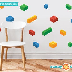May include: Reusable fabric decals of colorful building blocks in various sizes and colors, including red, blue, green, and yellow. The decals are shown on a white wall with a white chair in the foreground. The text "REUSABLE FABRIC DECALS" and "sunny decals" are visible in the image.