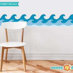 Puede incluir: Calcoman&iacute;as de pared de tela reutilizables con un borde de olas azules. Las calcoman&iacute;as se muestran en una pared blanca con una silla blanca delante de ellas. El texto "REUSABLE FABRIC DECALS" est&aacute; en la parte superior de la imagen. El texto "sunny decals" est&aacute; en la parte inferior de la imagen.