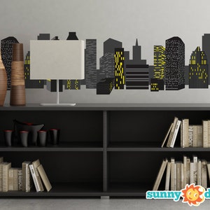 May include: Black bookshelf with two shelves, filled with books. A white lamp with a black base sits on top of the bookshelf. Two brown vases are on the left side of the bookshelf. A black and yellow cityscape decal is on the wall behind the bookshelf.