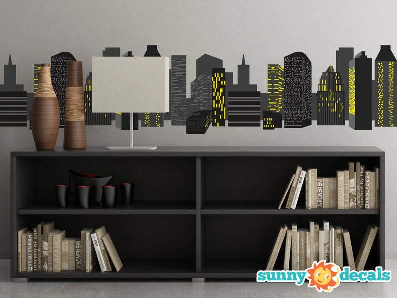 May include: Black bookshelf with two shelves, filled with books. A white lamp with a black base sits on top of the bookshelf. Two brown vases are on the left side of the bookshelf. A black and yellow cityscape decal is on the wall behind the bookshelf.
