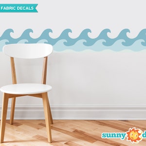 Puede incluir: Calcoman&iacute;as de pared de tela reutilizables con un patr&oacute;n repetitivo de olas azules del oc&eacute;ano. Las calcoman&iacute;as se muestran en una pared blanca con una silla blanca en primer plano. El texto "REUSABLE FABRIC DECALS" es visible en la esquina superior izquierda de la imagen. El texto "sunny decals" es visible en la esquina inferior derecha de la imagen.